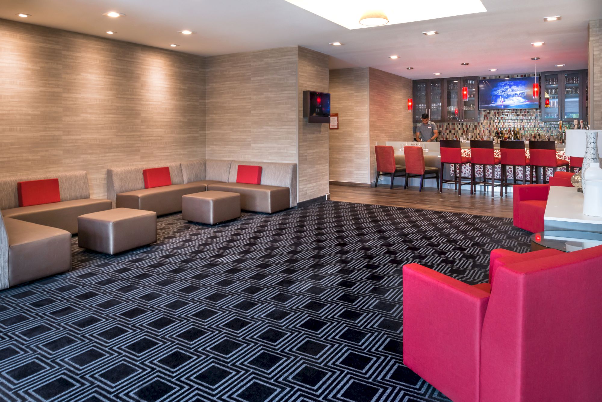 The image depicts a modern lounge area with patterned carpet, red and beige seating, and a bar setup in the background with red chairs.