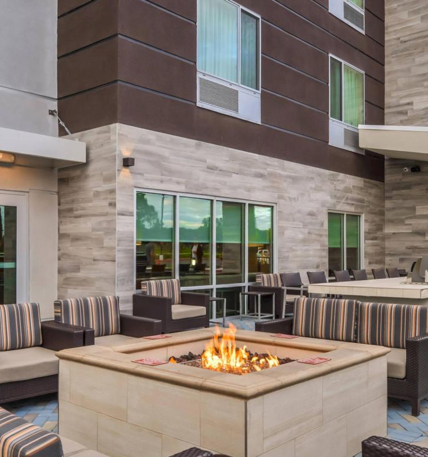 The image shows an outdoor patio area with a fire pit, surrounded by cushioned seating. There's also a BBQ grill station and a building in the background.