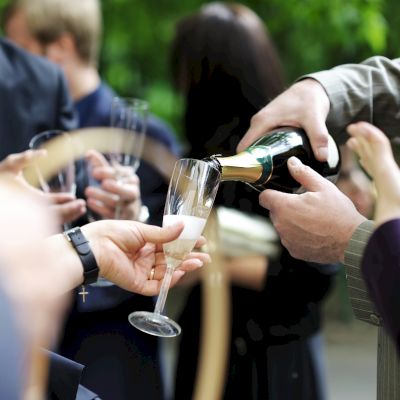 People in formal attire are gathered, with one person pouring champagne into a glass held by another, suggesting a celebratory event.