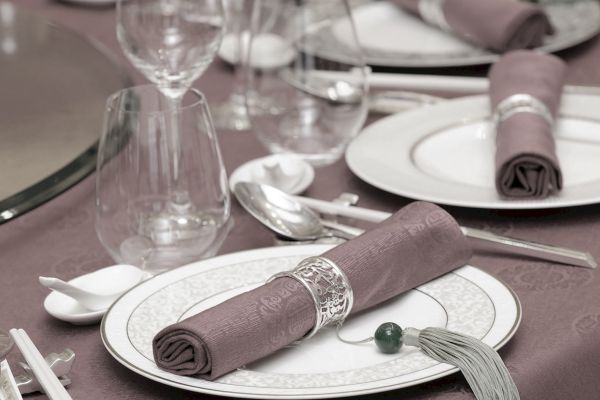 A formal dining table is set with plates, utensils, glasses, and neatly rolled napkins held by napkin rings on a mauve tablecloth.