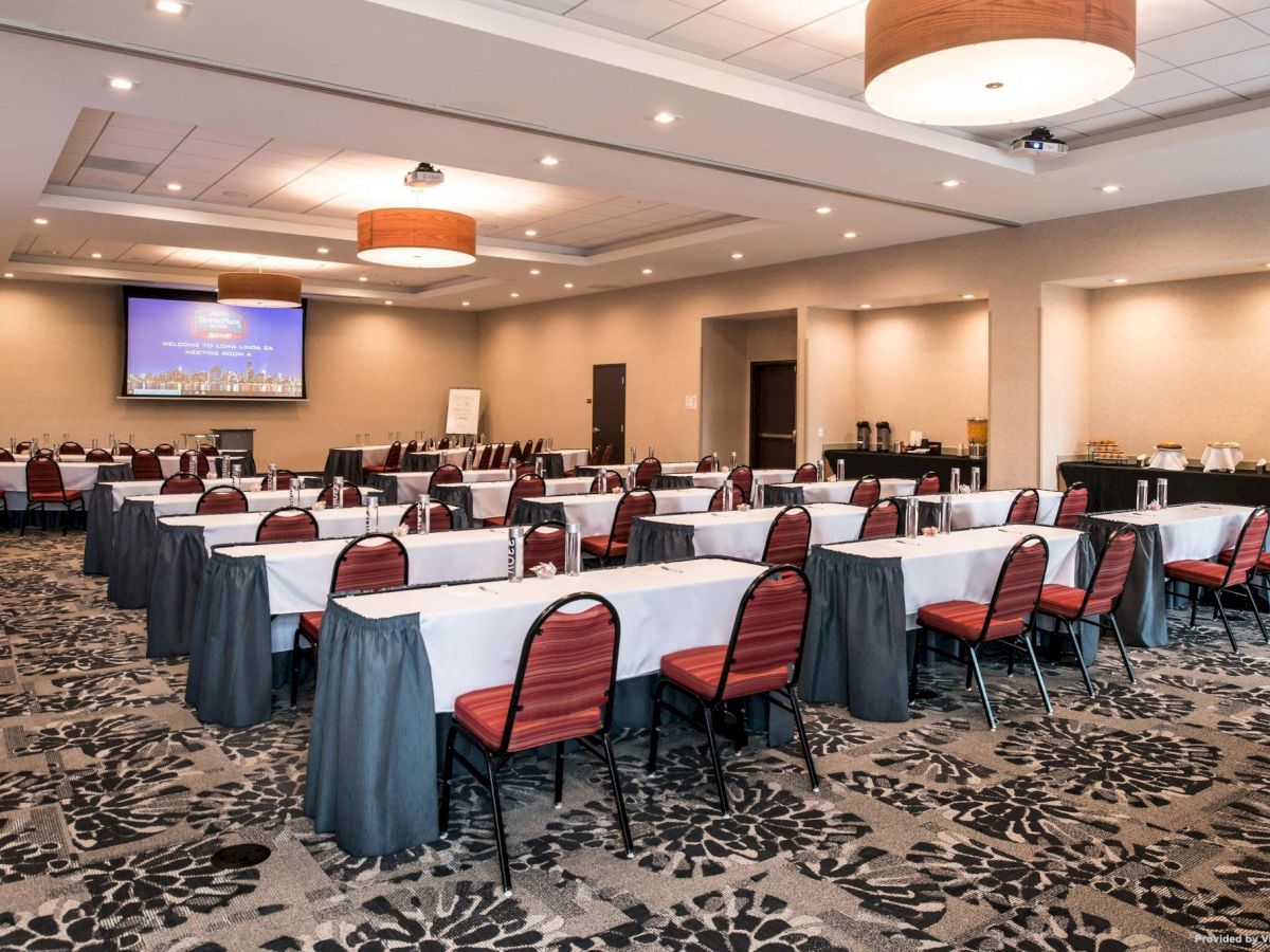 A conference room with tables and chairs arranged in rows facing a large screen. The room has a patterned carpet, ceiling lights, and a refreshment area.