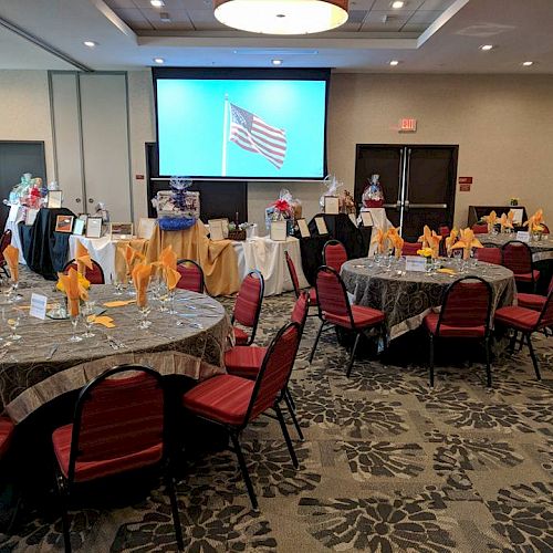 The image shows a decorated event space with round tables, red chairs, and place settings, featuring a screen displaying the U.S. flag at the front.