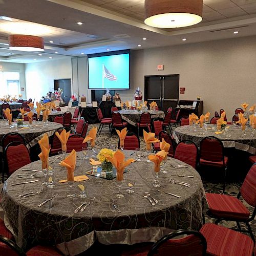 A banquet hall with round tables covered in gray tablecloths, set with yellow napkins, flowers, and dining utensils. A screen displays an image.