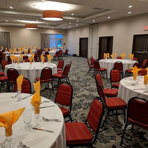 A banquet hall with round tables set with white tablecloths, red chairs, yellow napkins in glasses, and place settings, ready for an event or meeting.