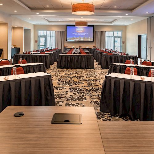 A conference room with rows of tables and chairs, projector screens displaying a cityscape image, and some water bottles and notepads on the tables.