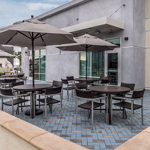 The image shows an outdoor dining area with round tables, chairs, and umbrellas on a paved patio next to a modern building.