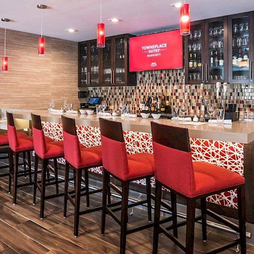 This image shows a modern bar area with red high chairs, a countertop with place settings, a tiled backsplash, and a display shelf with glasses and bottles.