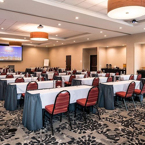 A spacious conference room with neatly arranged tables and chairs, a large screen displaying a presentation, and refreshments on a side table.