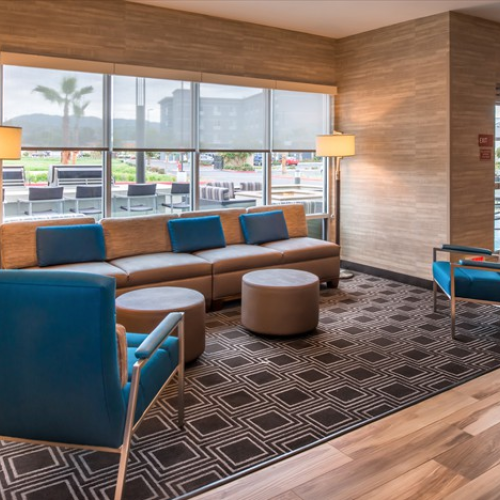 A modern waiting area with blue chairs, a grey sofa, two round ottomans, warm lighting, and large windows overlooking an outdoor area.