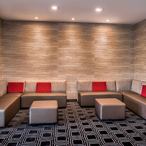 The image shows a modern lounge area with beige sectional seating, red cushions, two square ottomans, and a geometric patterned carpet, with subtle lighting.