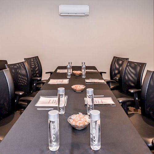 A modern conference room with black chairs, a long table set with water bottles, notepads, pens, and bowls of candy.