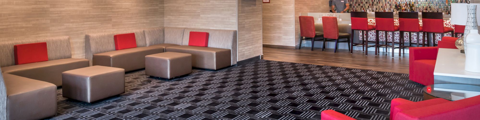 The image shows a modern lounge area with patterned carpet, red and gray seating, and a bar with high chairs and pendant lights in the background.