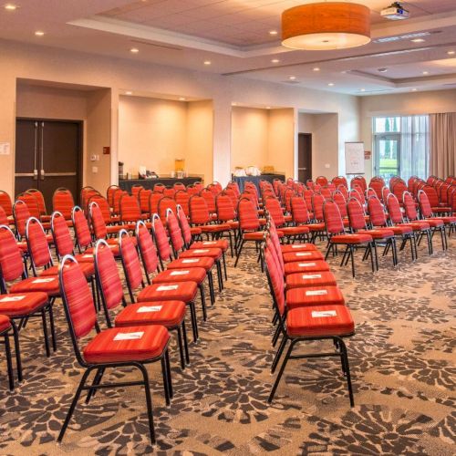 A conference room with red chairs arranged in rows, a podium, and a projector screen displaying a welcome message. The setup is ready for an event.