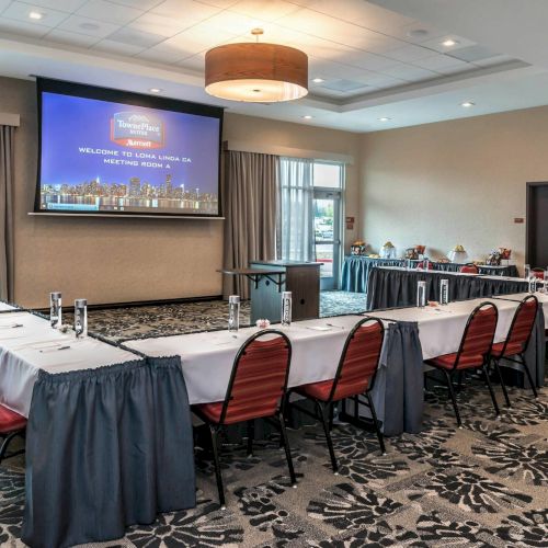 A conference room is set up with tables and chairs arranged in a U-shape, facing a projector screen displaying a welcome message.