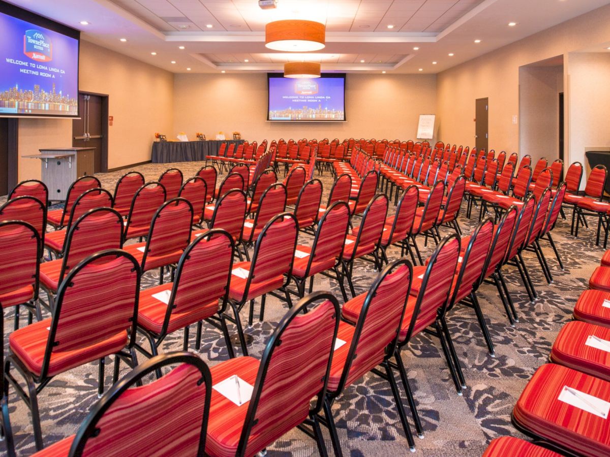 The image shows an empty conference room with rows of red chairs facing a stage area with presentation screens at the front of the room.