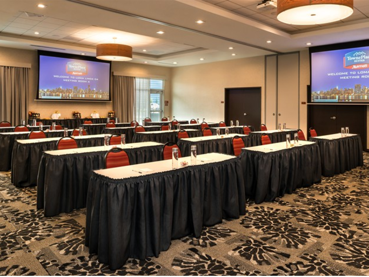 A conference room with rows of tables and chairs, two large screens with a welcome message, and water bottles set up on the tables.