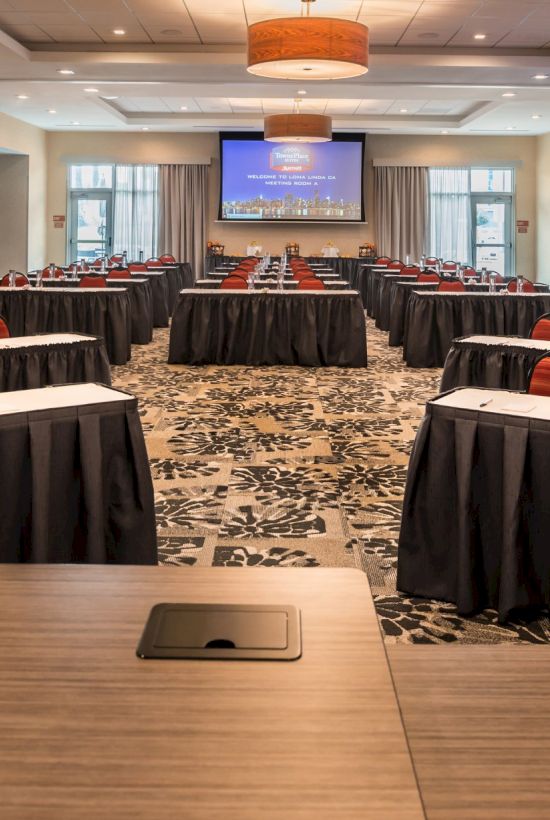 A conference room set up with rows of tables, chairs, and water bottles, featuring large presentation screens in a spacious, well-lit environment.