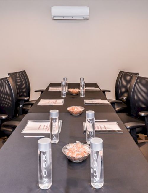 A modern conference room with a long table, black chairs, water bottles, documents, and bowls of candy, with an air conditioner above.