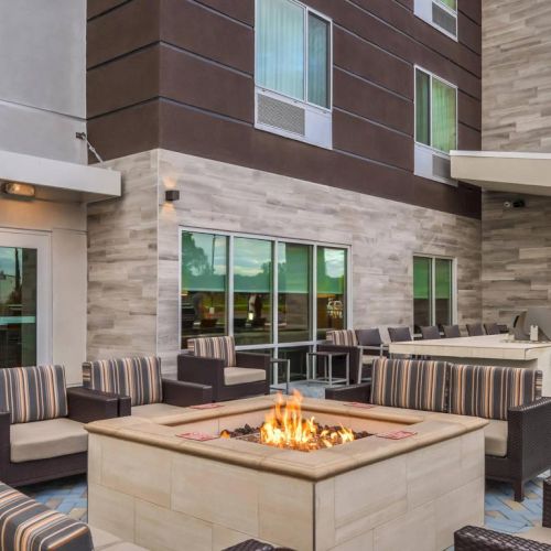 An outdoor seating area with a fire pit, surrounded by striped cushioned chairs, and a barbecue grill at the edge of a modern building.