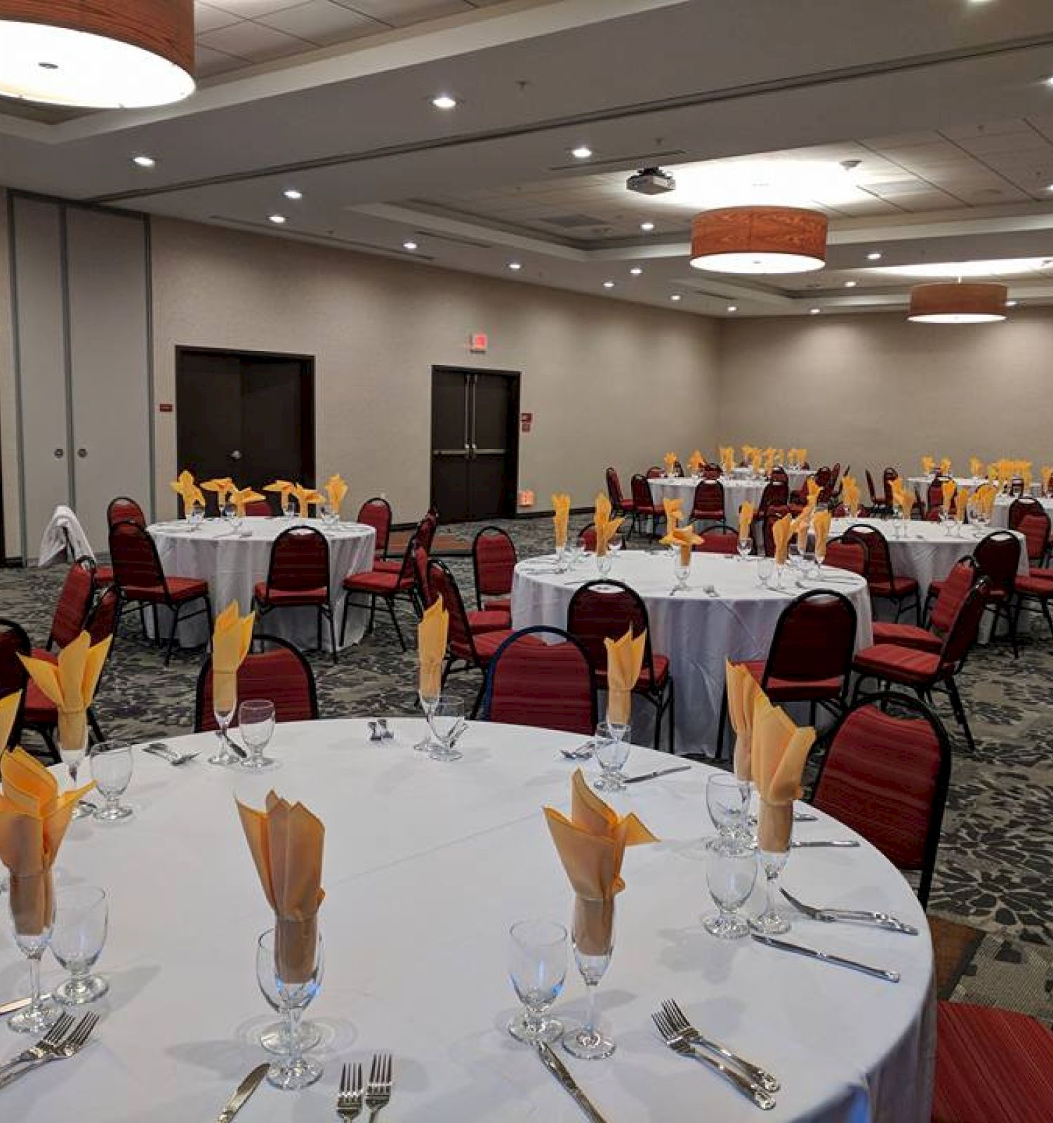 A banquet hall with round tables set for an event, featuring arranged cutlery, glasses, yellow napkins, and red upholstered chairs.