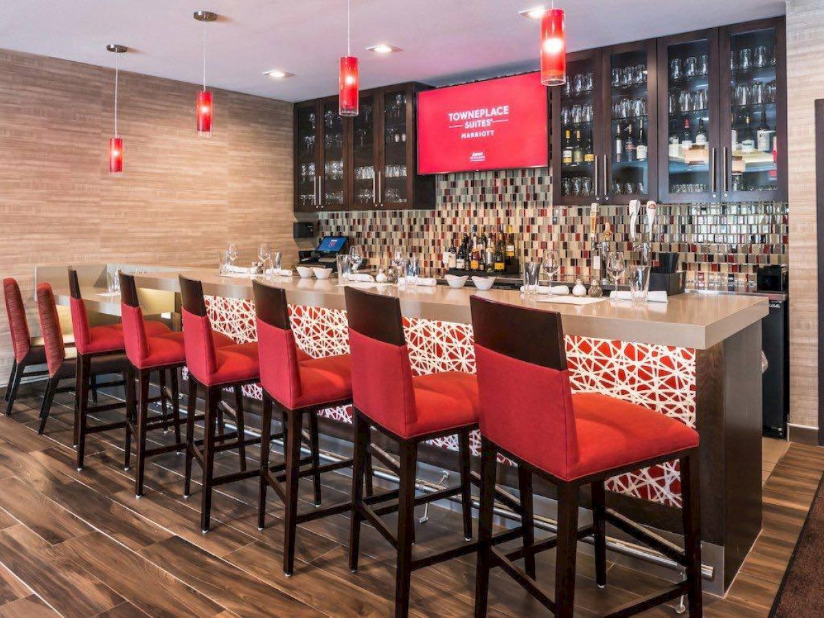 The image shows a modern bar with red stools, a countertop, a variety of bottles, and a display screen with text. The overall design is stylish.