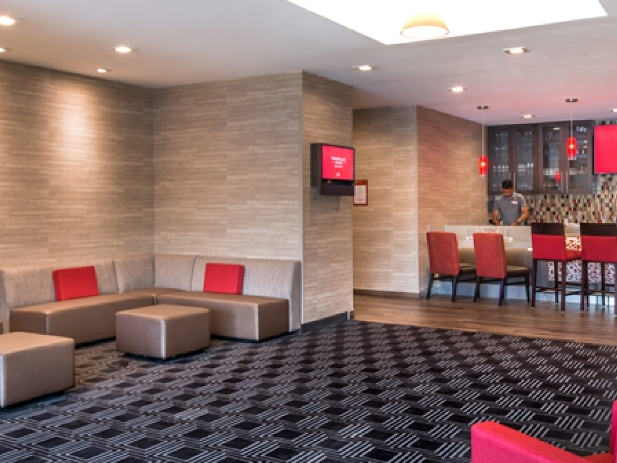 A modern lounge area with grey couches, red cushions, and a patterned carpet is adjacent to a bar counter with red chairs and a bartender.