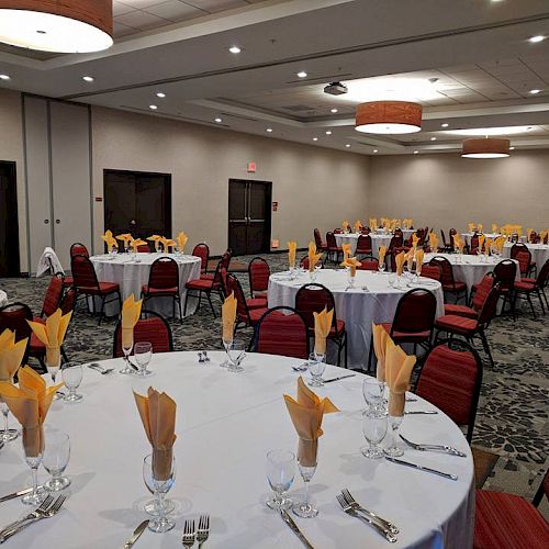 A banquet hall is set up with round tables, red chairs, and table settings, including orange napkins, glassware, and silverware, in a well-lit room.