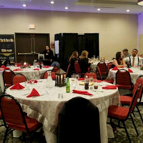 People are gathered in a banquet hall with round tables and red chairs, engaging in conversation, enjoying food and drinks, and socializing.