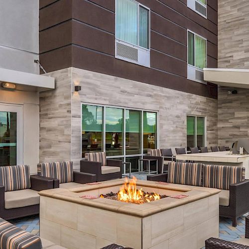 An outdoor patio with cushioned seating around a fire pit, a grill area, and a modern building in the background with large windows and multiple floors.