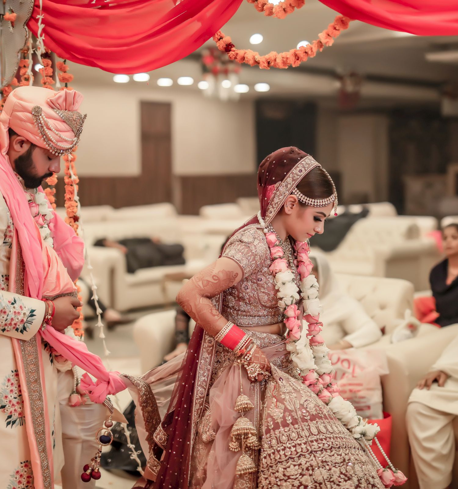 A bride and groom wear traditional attire in a decorated setting with several seated guests, suggesting a wedding ceremony.