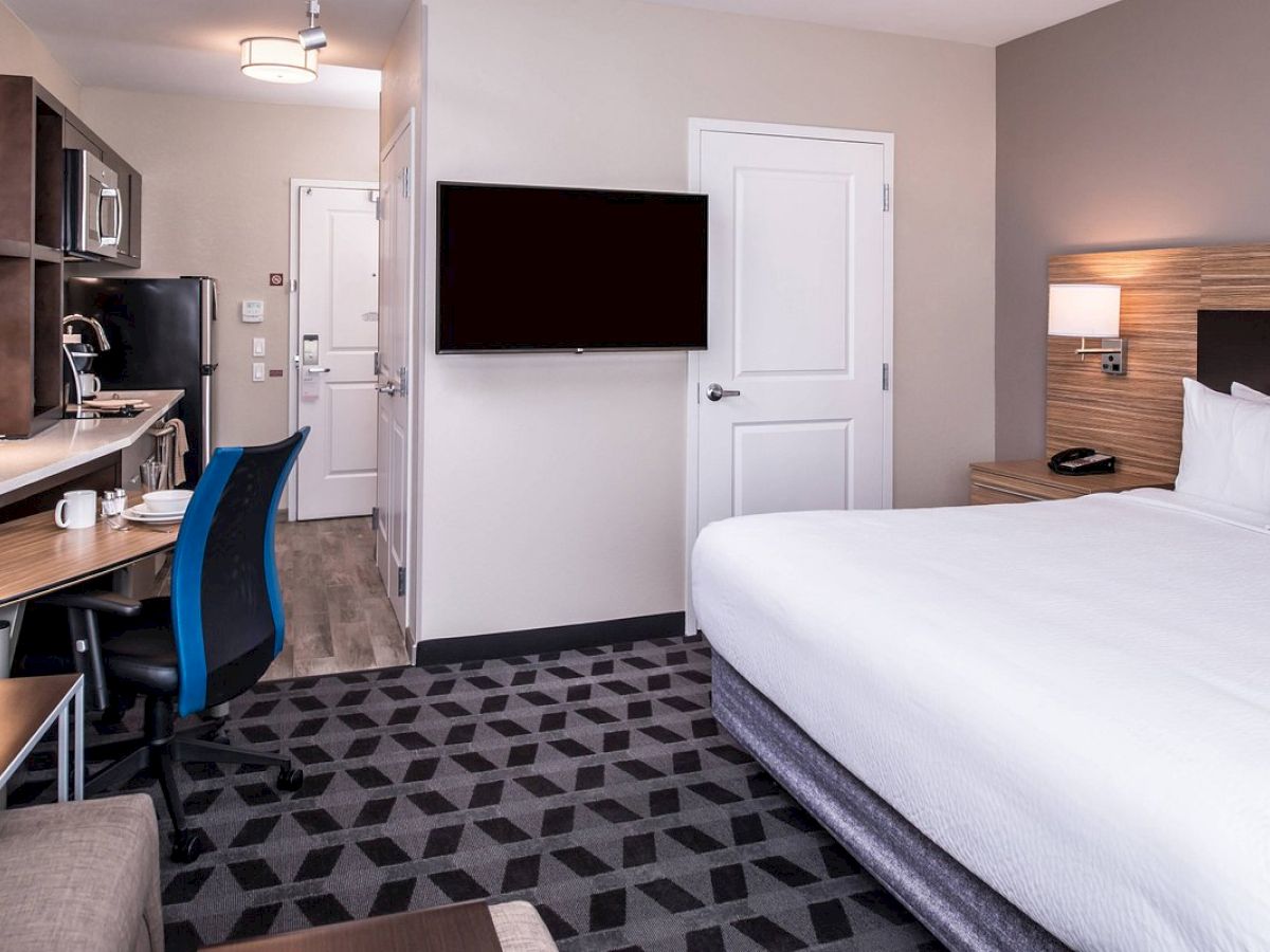 A modern hotel room featuring a large bed, a wall-mounted TV, a desk with a chair, and a kitchenette with appliances and a dining area.