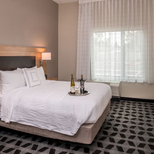 A neatly made bed in a hotel room with two bottles of wine on a tray, surrounded by modern furniture near a large window with sheer curtains.