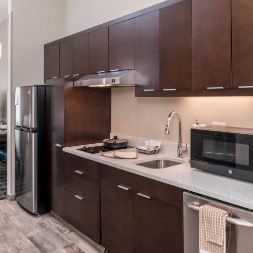 A modern kitchen with dark wood cabinets, stainless steel appliances, and a small dining area in the background featuring a TV and chairs.