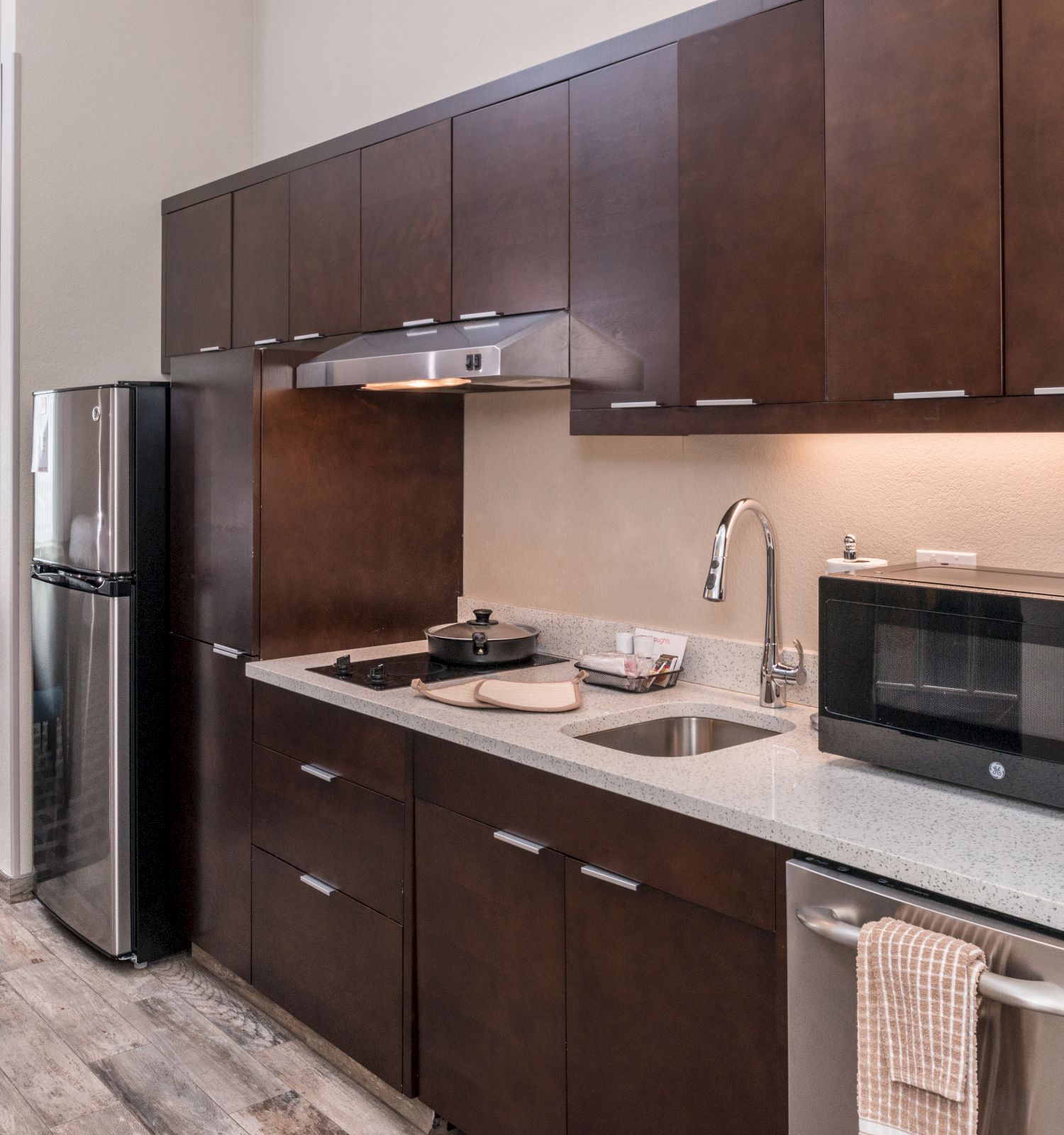 A modern kitchen with dark wood cabinets, a microwave, coffee maker, sink, stove, and refrigerator; a dining area is visible in the background.