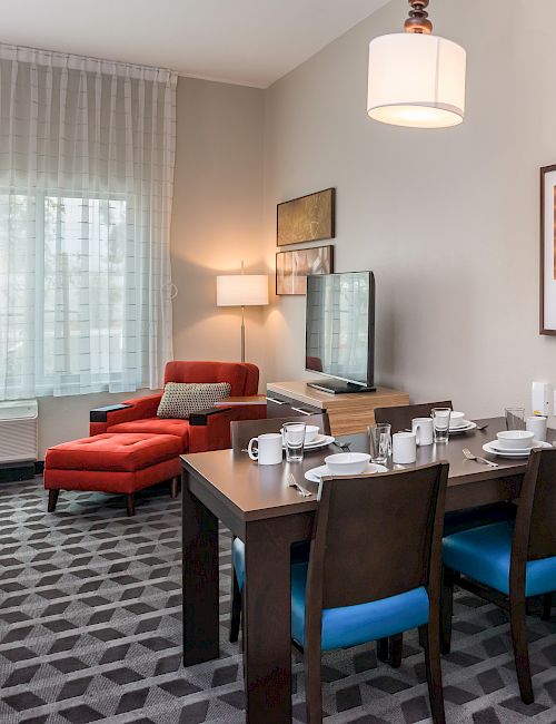 This image shows a modern kitchen and living area with a stainless steel fridge, dining table, blue chairs, and a lounge area with a red chair.