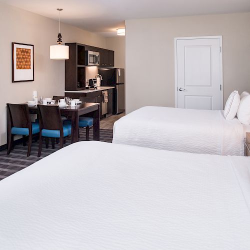 The image shows a hotel room with two white-sheeted beds, a kitchenette, a dining table with chairs, a TV, and soft lighting for a cozy atmosphere.