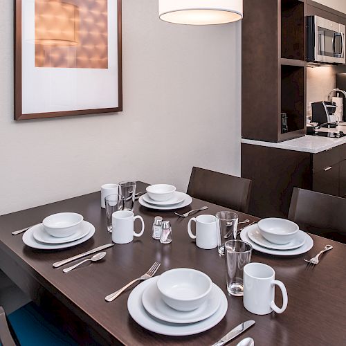 A modern kitchen and dining area with dark wood furniture, fully set dining table for six, microwave, stove, refrigerator, and a framed picture on the wall.